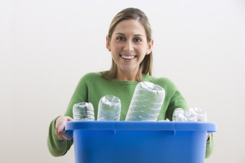 A young woman with light brown hair wearing a bright green sweater, smiling and holding a blue plastic recycling bin filled with clear plastic bottles of various sizes. The bottles appear to be household drinking containers, with some standing upright and others leaning against each other within the bin. The background is plain and white, emphasizing the person's cheerful expression and the focus on the recycling activity. This image relates to waste management and recycling services that could be offered by Commercial Waste Hackbridge, a company serving the Hackbridge area in London. The scene is well-lit, with natural or studio lighting highlighting the smooth textures of the plastic bottles and the vibrant color of the sweater, conveying a positive and environmentally conscious message about rubbish collection and recycling efforts in the local community.