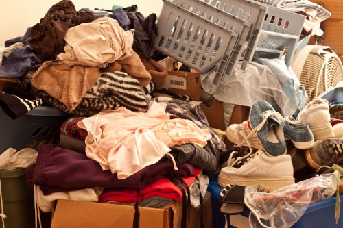 A large pile of mixed household clothing, fabrics, and textiles in various colours and materials, including knitted, woven, and cotton items, piled on a surface with some hanging over the edge. Among the clothes are shoes, with a pair of white sneakers visible, and plastic bags partially containing more items. In the background, a plastic laundry basket is semi-tilted, with more garments and items stacked behind it, and a cardboard box partially visible at the bottom of the heap. The scene appears to be set in a domestic environment, possibly on a table or the floor, with natural lighting illuminating the cluttered arrangement, indicative of a decluttering or rubbish removal process, related to services like those provided by Commercial Waste Hackbridge in the local area near postcodes in the Hackbridge region.