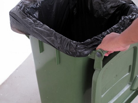 A close-up view of a green plastic waste bin with a black plastic liner inside, situated on a grey pavement surface. A person's hand is gripping the bin's handle, preparing to move or empty it. The bin is positioned outdoors on a driveway or street area, with natural lighting illuminating the scene. The environment suggests a typical setting for rubbish collection or waste removal services, in a residential or commercial area possibly within Hackbridge or nearby in the postcode region. The image emphasizes the practical aspect of rubbish disposal, aligning with commercial waste removal activities undertaken by companies such as Commercial Waste Hackbridge, focusing on efficient and tidy rubbish management in London suburbs.