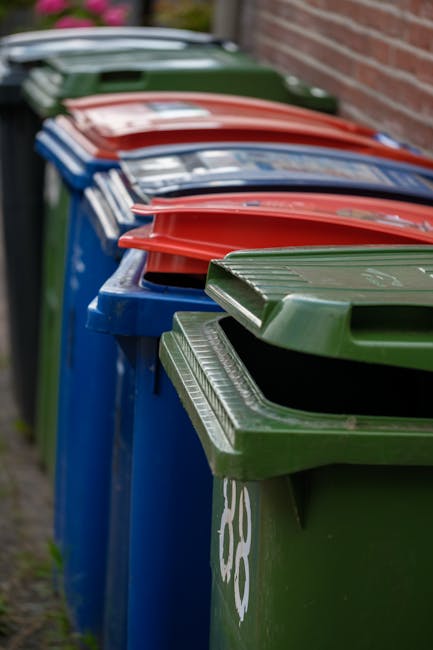 A large green plastic waste skip positioned on a paved surface, placed against a black brick wall. The skip is filled with red and black flexible vacuum hose bags, with visible text on the bags indica